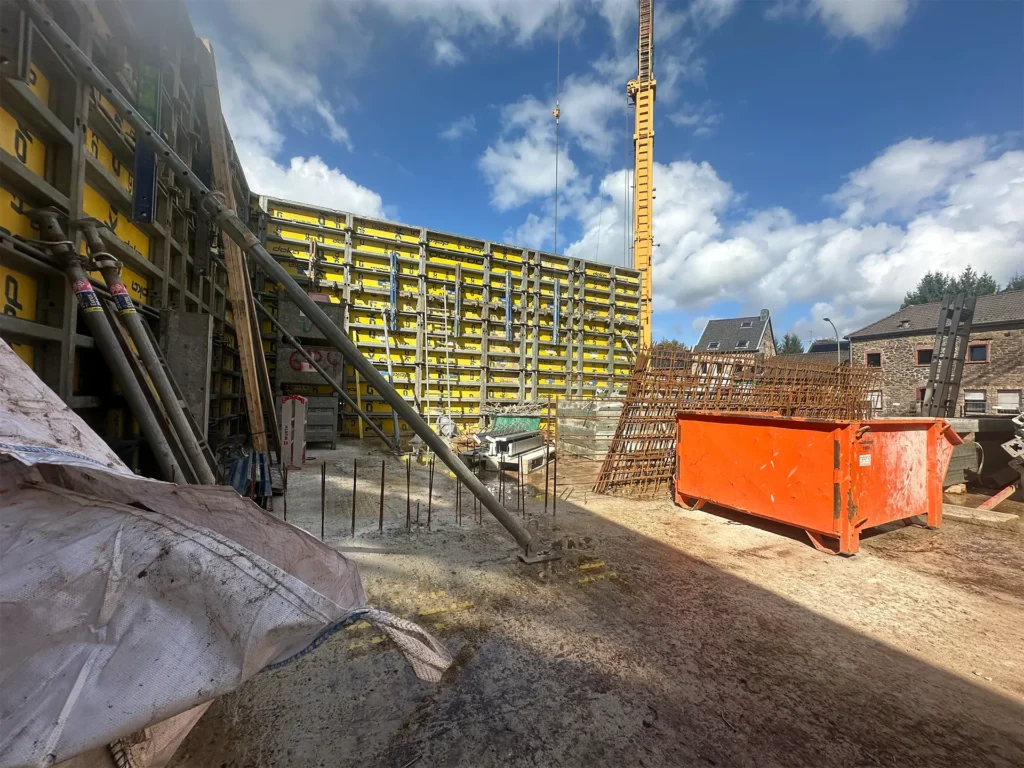 Innenansicht der Baustelle: Gelbe Schalungselemente bilden eine großflächige Wand, davor liegen Bewehrungsgitter, Bauholz und eine rote Mulde für Schutt. Links und rechts sind Baumaterialien und Werkzeuge verteilt, im Hintergrund erkennt man Wohnhäuser.