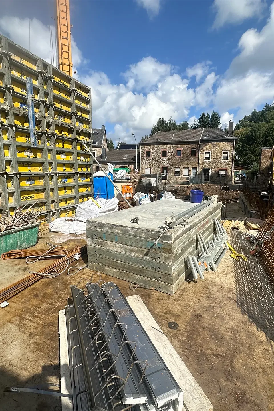 Blick in den Innenbereich der Baustelle mit großflächigen gelben Schalungselementen. Auf dem Boden liegen Holzbohlen, Eisenstangen und Bauzubehör. Rechts lehnt ein Gitter aus Bewehrungsstahl an der Wand, im Hintergrund sind Wohnhäuser und ein Bagger zu sehen.
