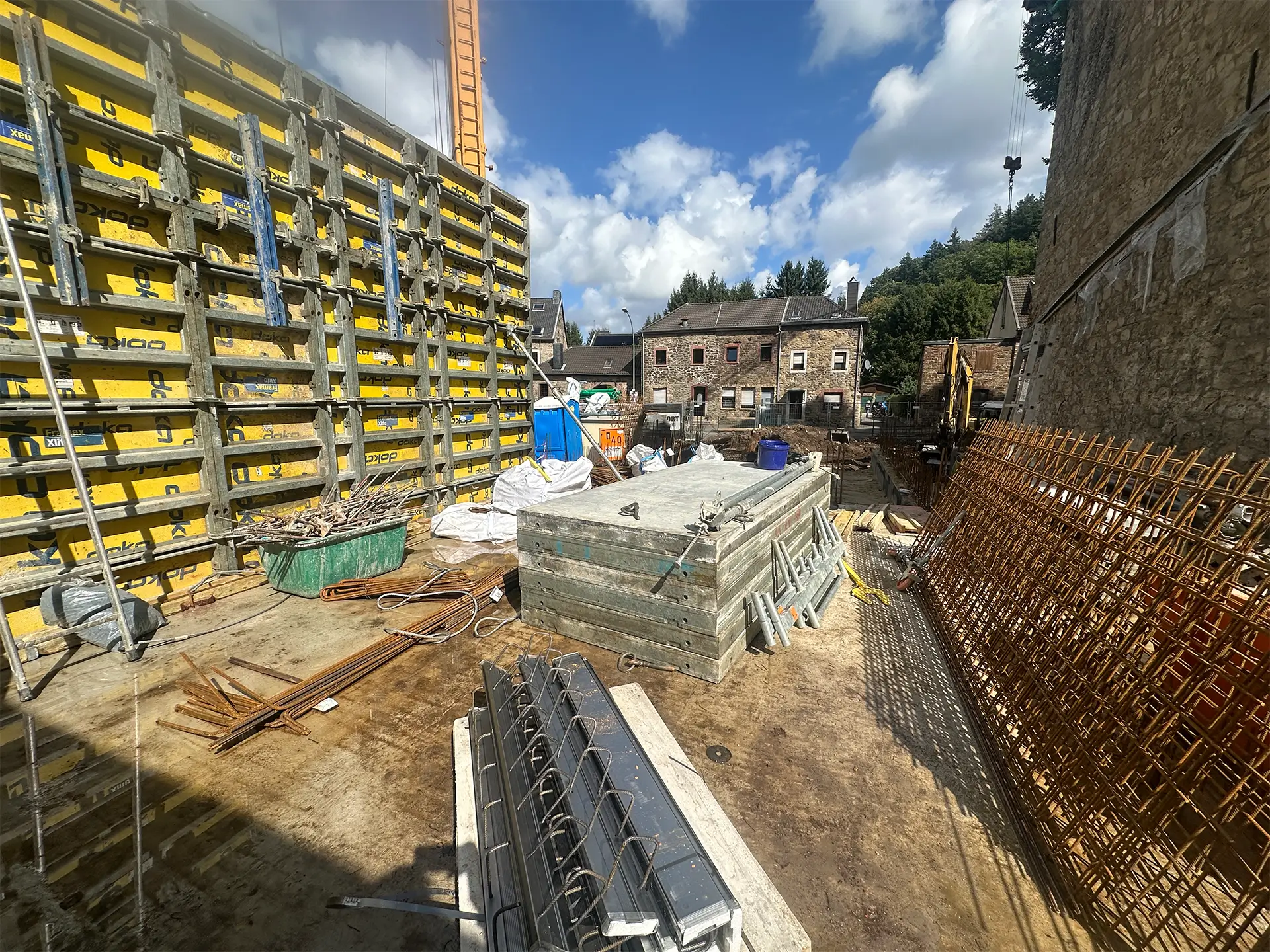 Blick in den Innenbereich der Baustelle mit großflächigen gelben Schalungselementen. Auf dem Boden liegen Holzbohlen, Eisenstangen und Bauzubehör. Rechts lehnt ein Gitter aus Bewehrungsstahl an der Wand, im Hintergrund sind Wohnhäuser und ein Bagger zu sehen.