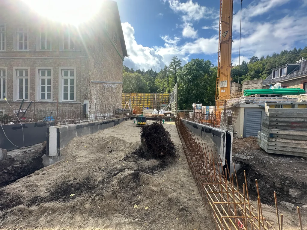 Blick auf die Baustelle neben einem historischen Backsteingebäude, wo die Fundamente und Bewehrungen für den Neubau der Kita vorbereitet sind. Im Vordergrund liegt aufgeschütteter Boden, rechts und links ragen Stahlarmierungen empor. Ein Kran und Baumaterialien sind im Hintergrund sichtbar.