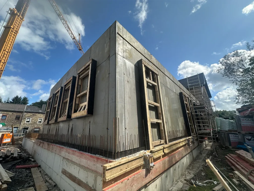 Nahaufnahme einer neu errichteten Betonwand des Kita-Neubaus. Mehrere Holzrahmen markieren zukünftige Fensteröffnungen. Im Hintergrund ist das historische Gebäude eingerüstet, daneben stapeln sich Baumaterialien. Ein Kran ragt in den blauen Himmel.