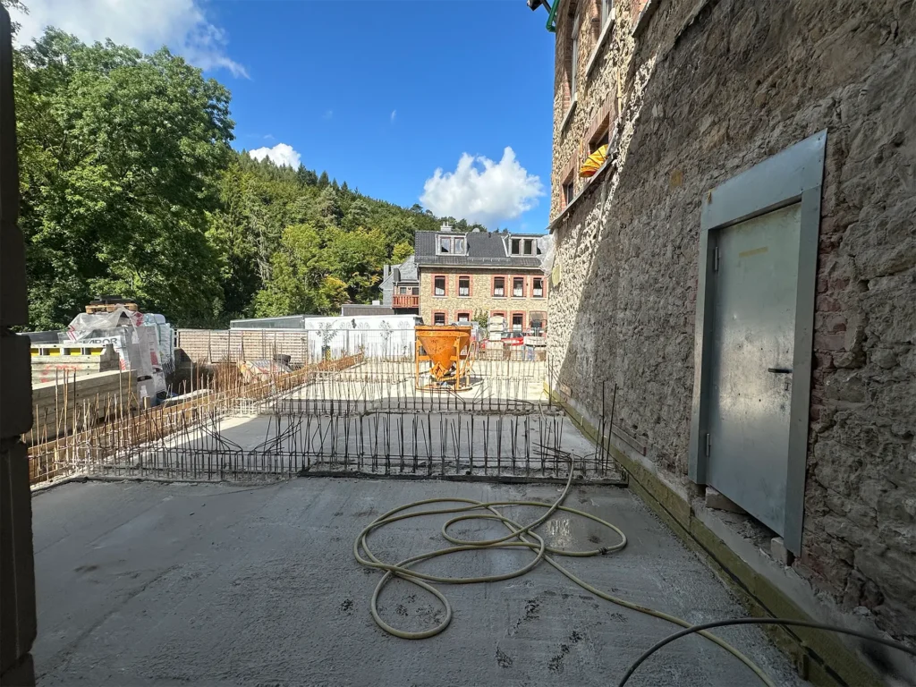 Bauarbeiten im Außenbereich mit neuer Betonplatte, Baustahl und Blick auf historische Gebäude sowie die grüne Umgebung