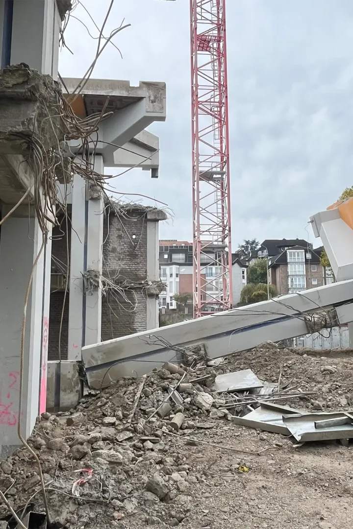 Teilweise abgerissenes Betongebäude mit freiliegenden Stahlarmierungen, umgeben von Schutt und Trümmern. Im Hintergrund ist ein roter Baukran zu sehen.