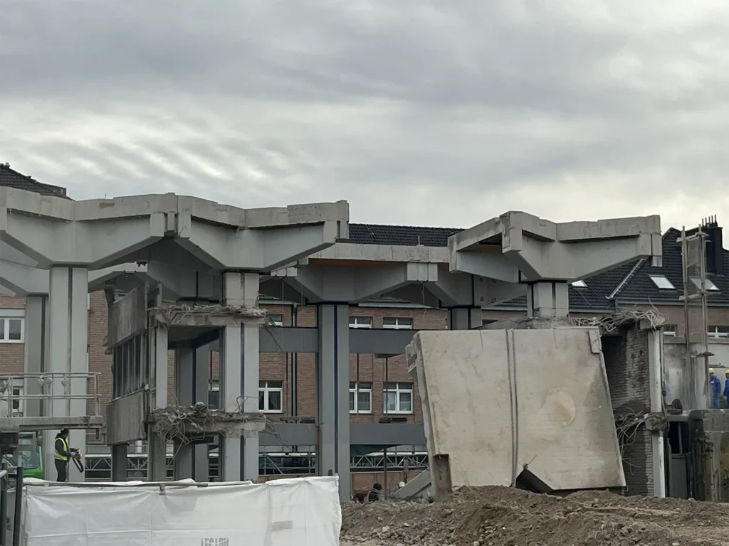 Ansicht eines Rohbaus während des Abrisses, bei dem große Betonträger und Wände bereits entfernt wurden. Vor den Gebäuderesten stehen Arbeiter und Baugerät.