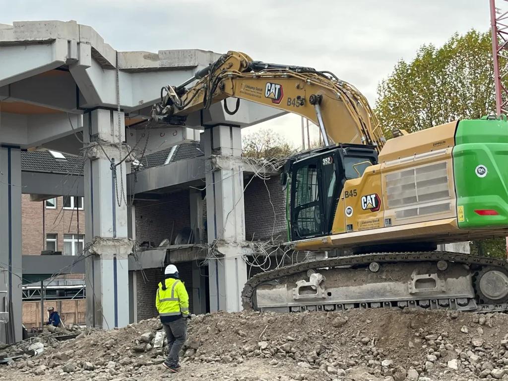 Bagger mit Abbruchzange arbeitet an der Fassade eines Betongebäudes. Ein Bauarbeiter in gelber Warnjacke beobachtet den Rückbau auf der Baustelle.