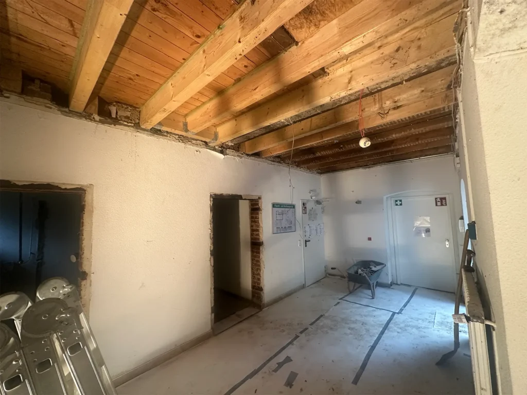 Innenaufnahme eines Gebäudes während der Sanierung – offene Holzdecke mit neuen Balken, entfernte Türrahmen und Baumaterial im Raum.