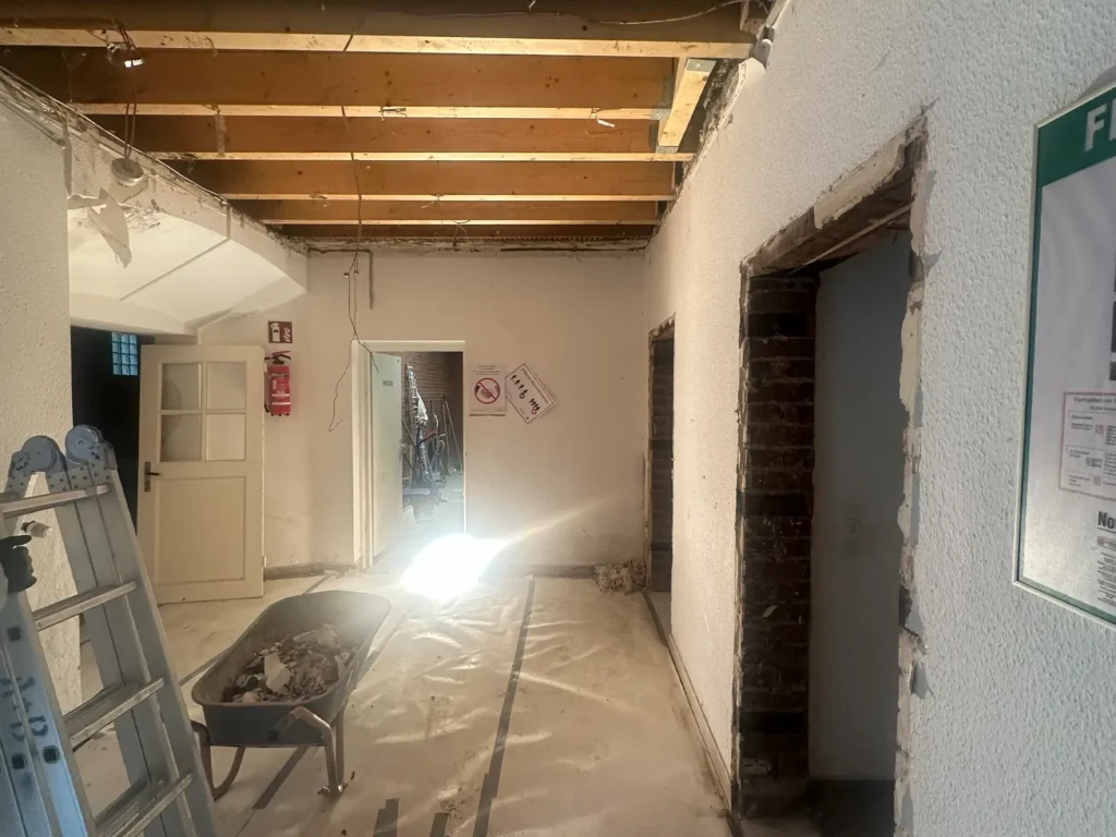 Blick durch einen sanierten Flur mit freigelegten Türöffnungen und neuer Holzdecke, im Vordergrund eine Schubkarre mit Bauschutt.