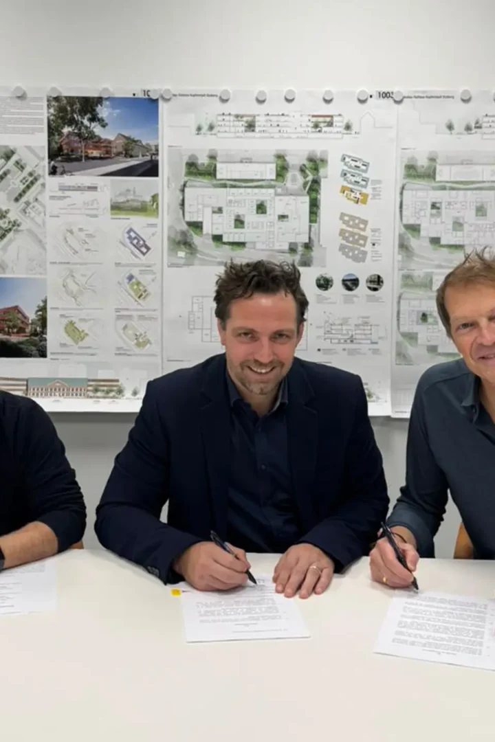 Drei Männer sitzen an einem Tisch und unterzeichnen Verträge, im Hintergrund hängen Architekturpläne an der Wand.