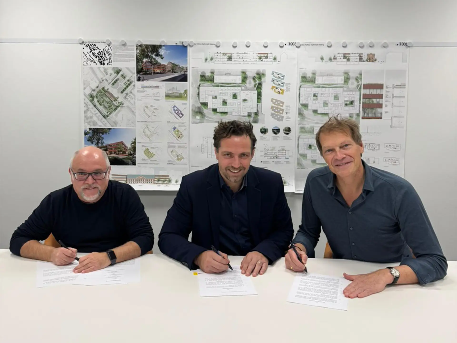 Drei Männer sitzen an einem Tisch und unterzeichnen Verträge, im Hintergrund hängen Architekturpläne an der Wand.