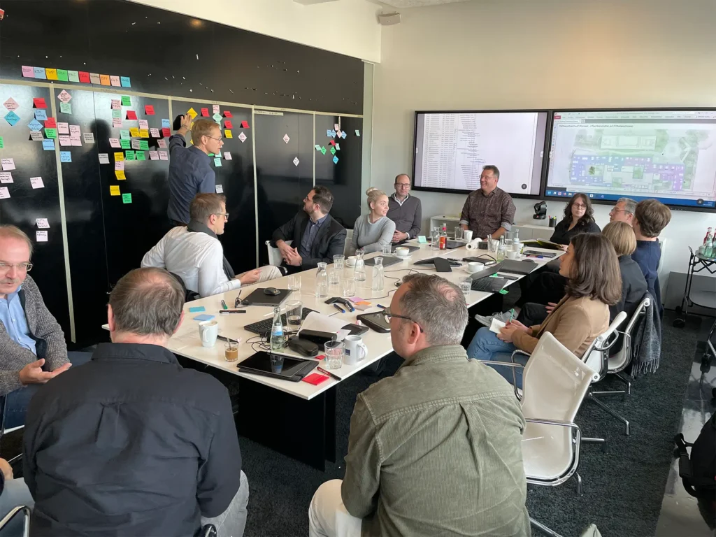 Teilnehmende eines Fachplaner-Workshops sitzen an einem Konferenztisch, während Inhalte an einer mit Haftnotizen belegten Wand diskutiert werden.