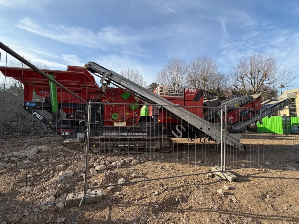 Große mobile Brechanlage auf einer abgesperrten Baustelle, die Bauschutt und Steine verarbeitet; Förderbänder und Maschinenkomponenten sind deutlich sichtbar.