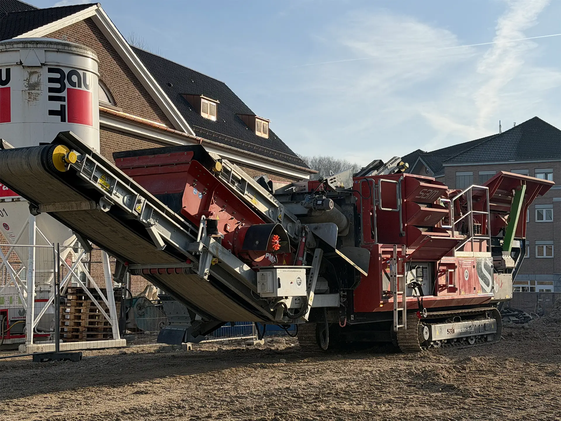Mobile Brech- und Siebanlage im Einsatz auf einer Baustelle zwischen Wohngebäuden, mit Förderband zur Materialaufbereitung und Schotterboden im Vordergrund.