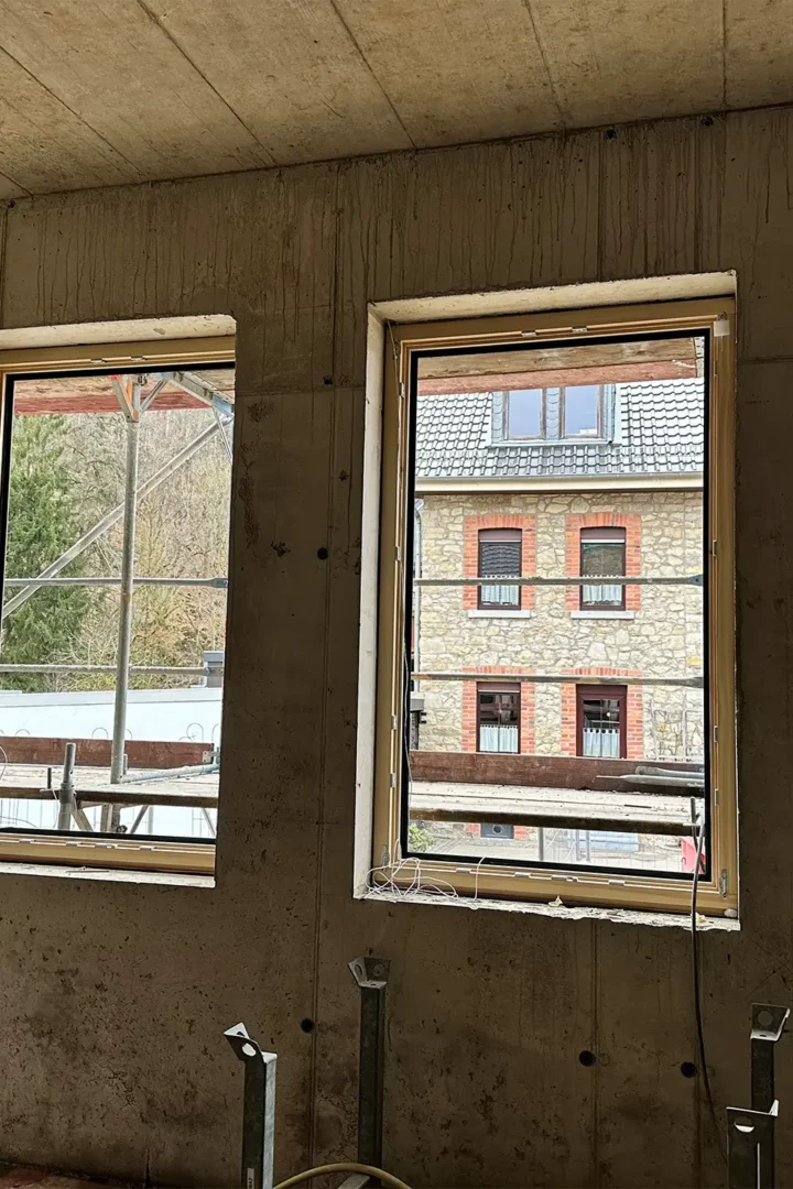 Neu eingesetzte Holzfenster im Rohbau des Anbaus; Blick von innen durch mehrere Fensteröffnungen auf das bestehende Gebäude und das Baugerüst.