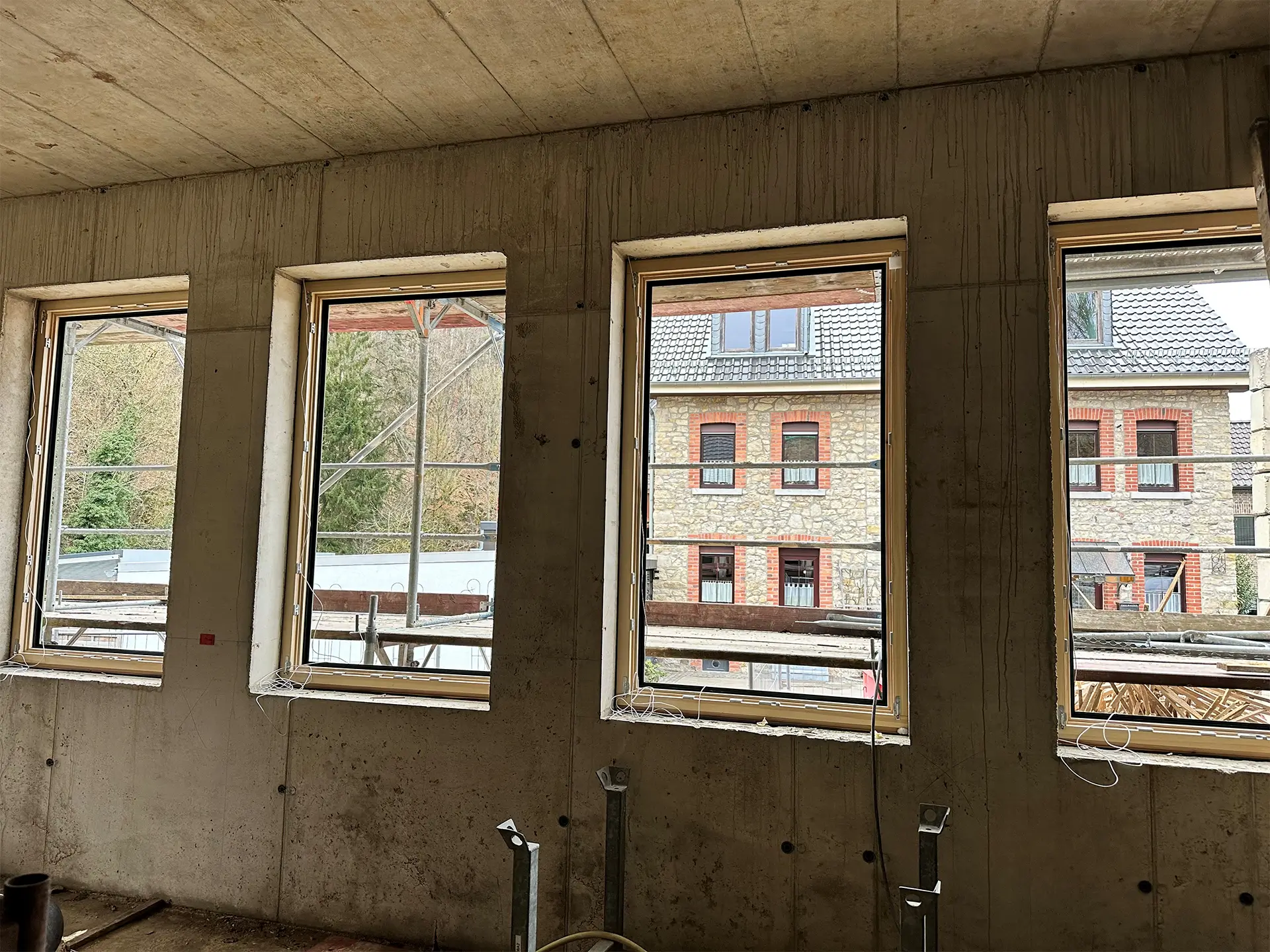 Neu eingesetzte Holzfenster im Rohbau des Anbaus; Blick von innen durch mehrere Fensteröffnungen auf das bestehende Gebäude und das Baugerüst.