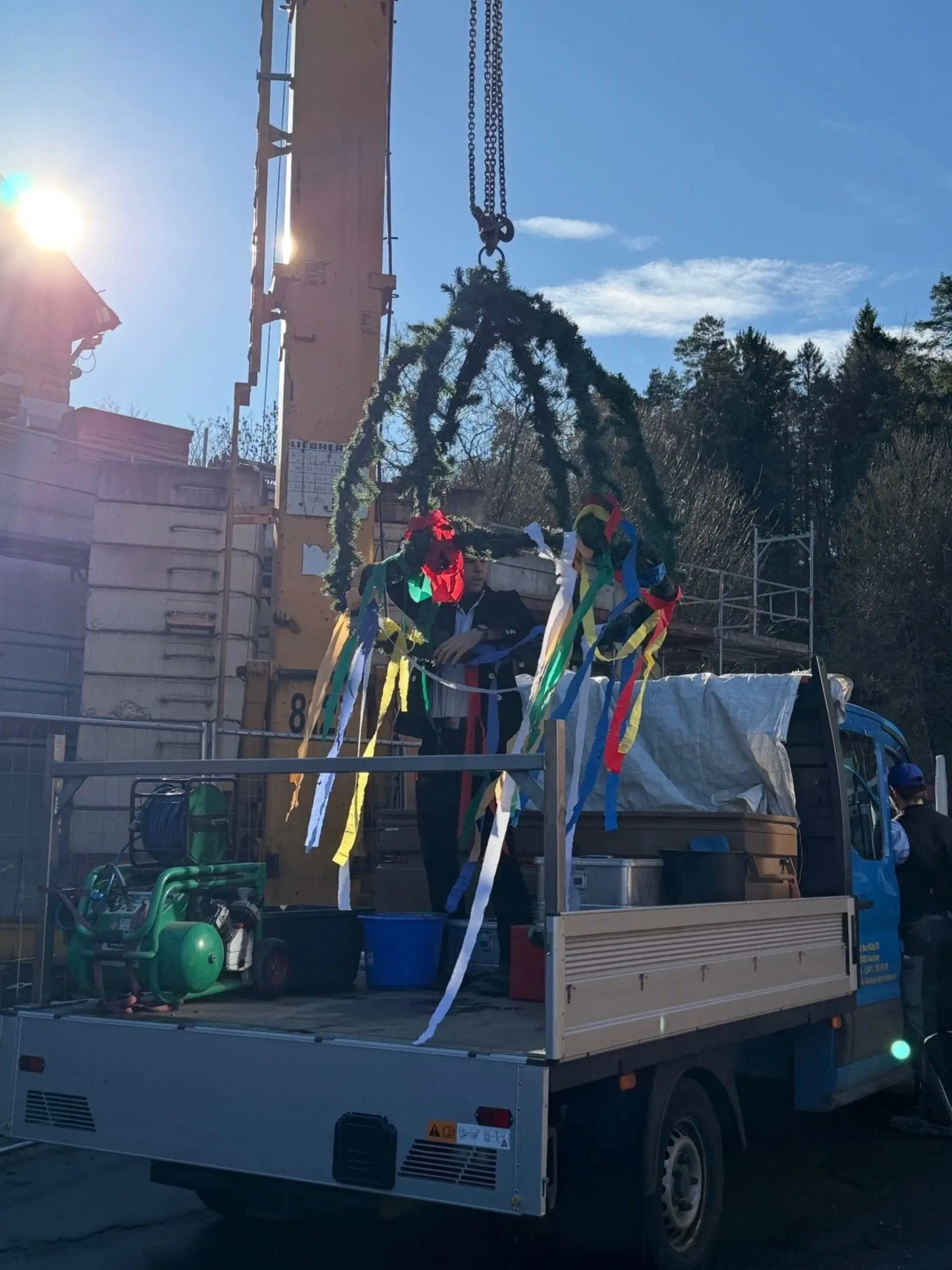 Eine Person steht auf der Ladefläche eines LKWs und richtet einen mit bunten Bändern geschmückten Richtkranz aus, der an einem Baukran hängt.