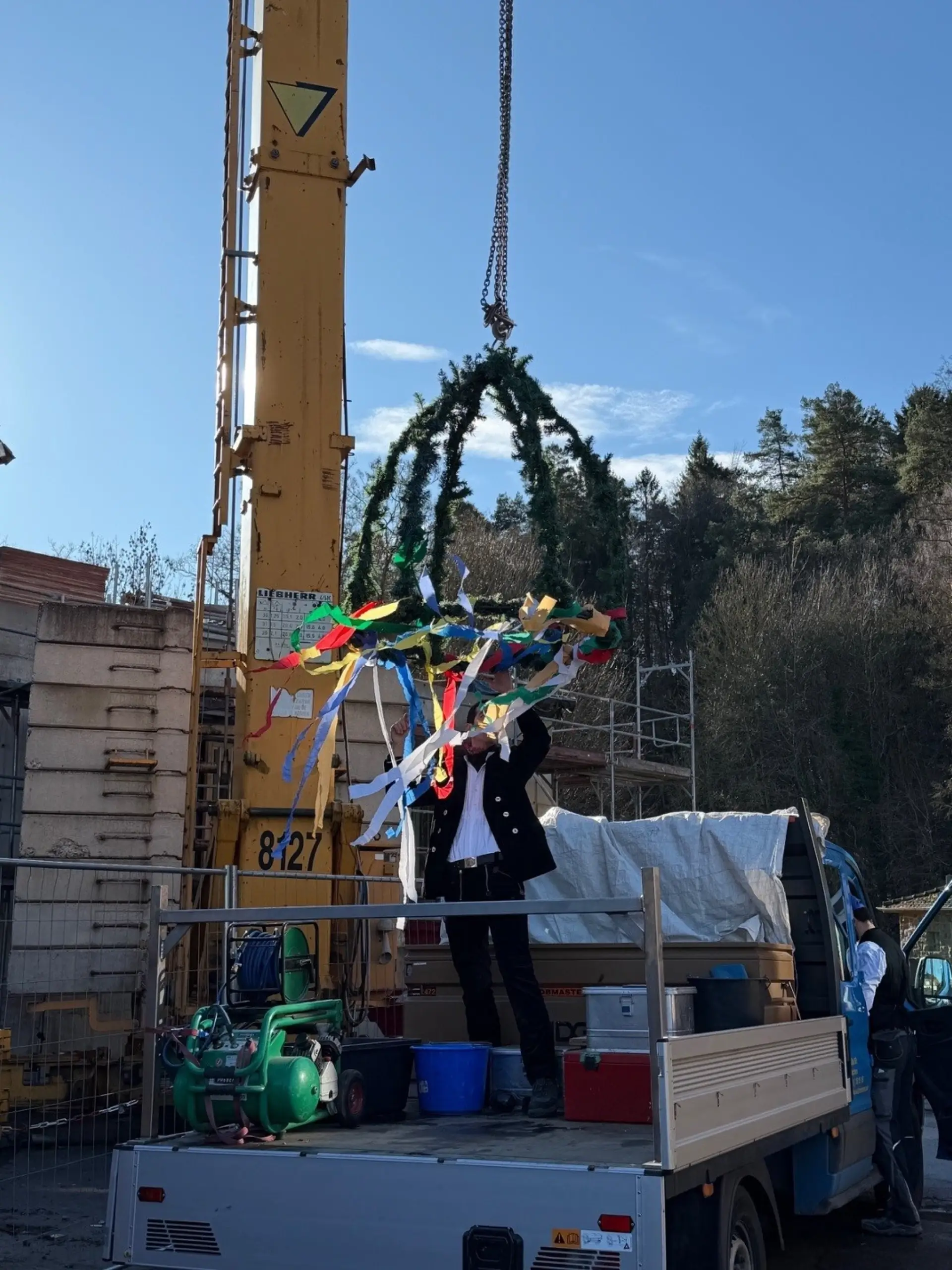 Der Richtkranz wird von einem Baukran angehoben, während eine Person darunter auf der Ladefläche eines LKWs steht.