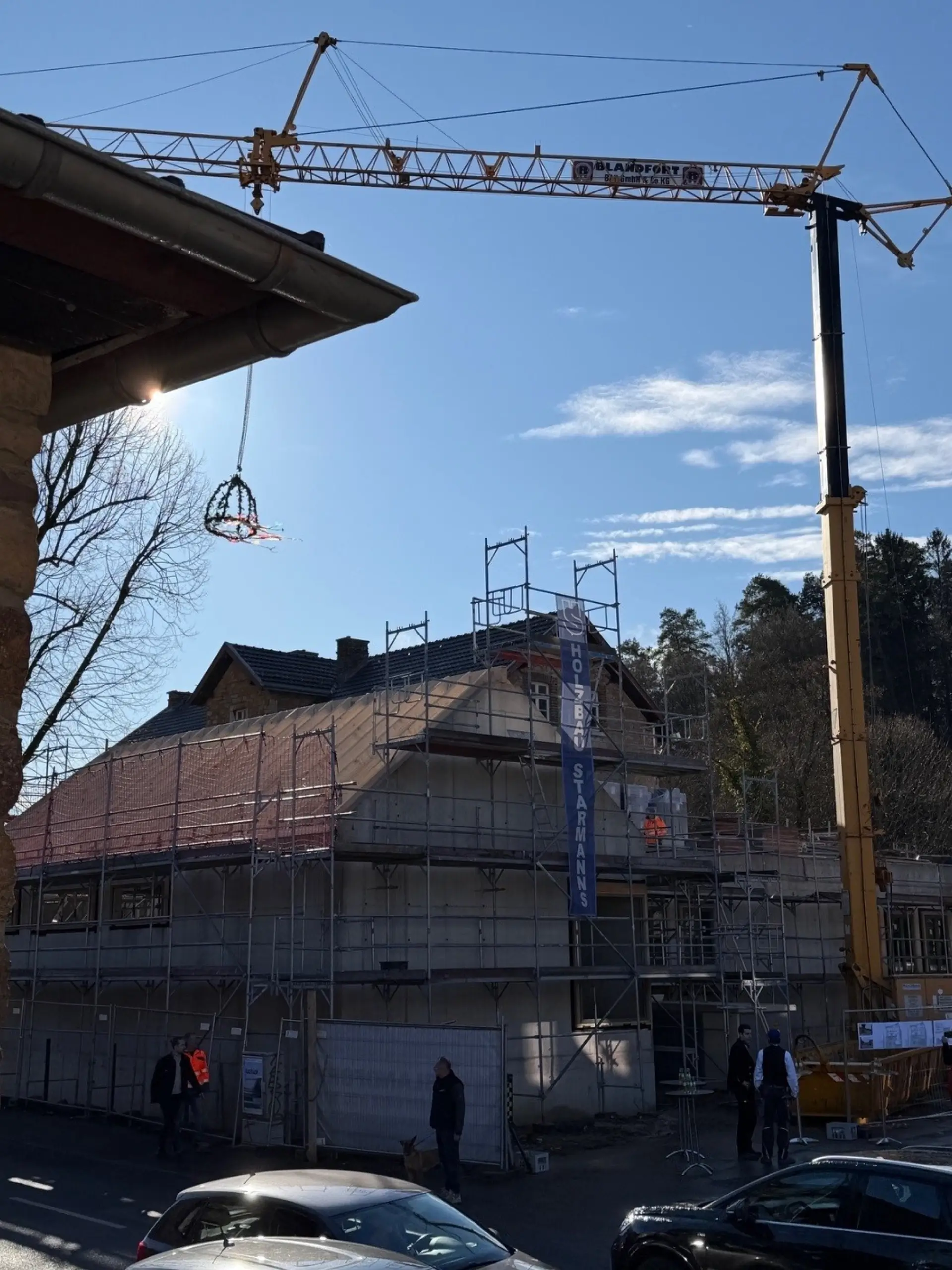 Ein Baukran spannt sich über ein eingerüstetes Gebäude, während ein Richtkranz über dem Dach schwebt.