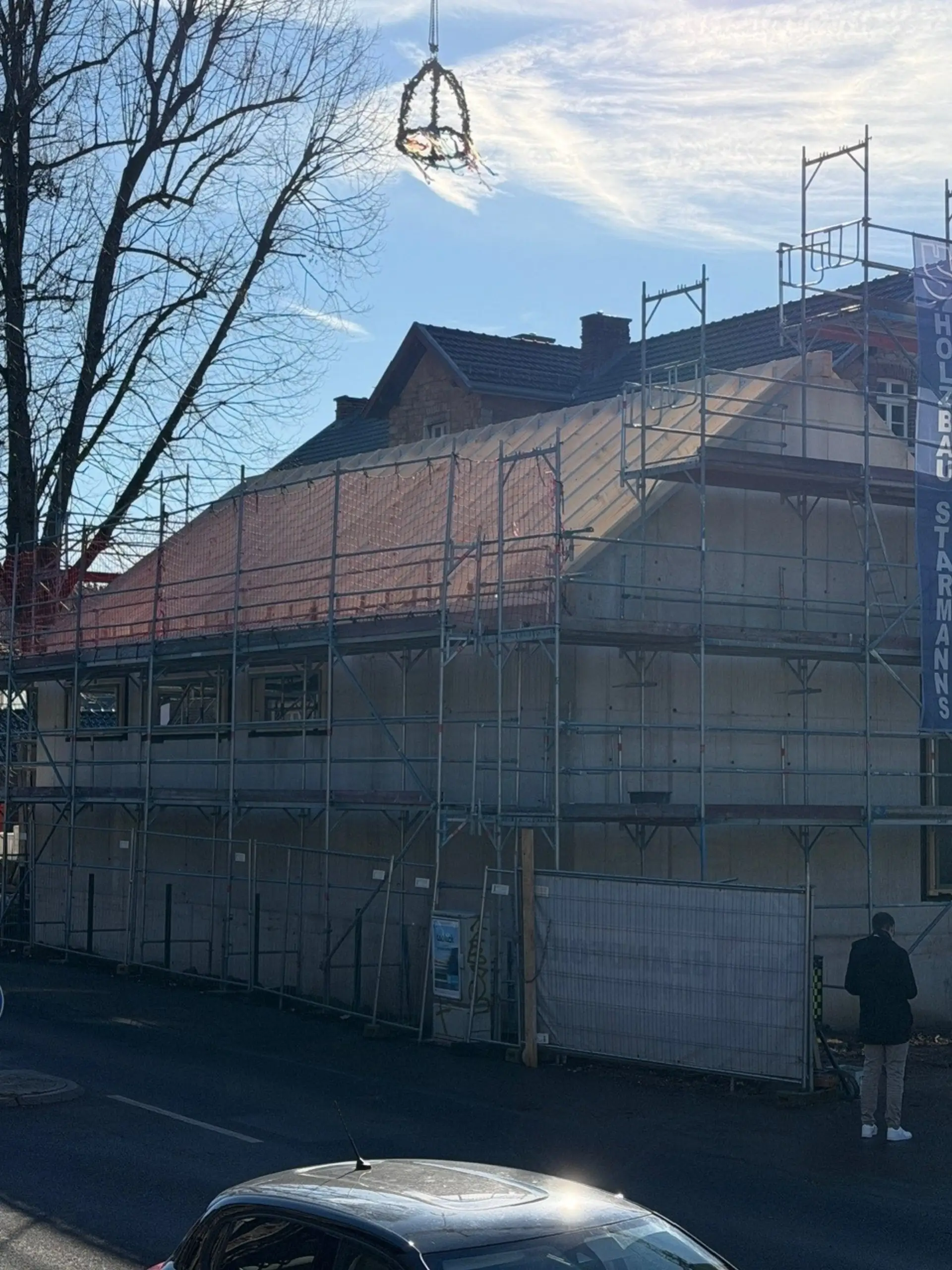 Der Richtkranz hängt hoch über einem eingerüsteten Gebäude mit teilweise gedecktem Dach vor blauem Himmel.