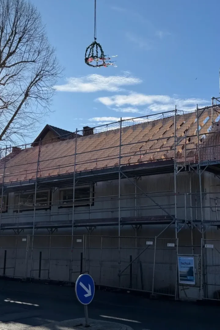 Weitaufnahme einer Baustelle mit eingerüstetem Gebäude, Baukran und schwebendem Richtkranz über dem Dach.