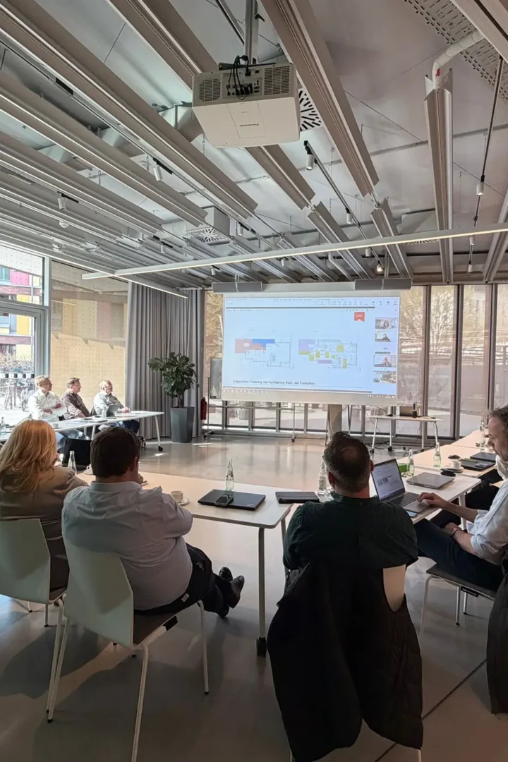 Gruppe von Teilnehmenden sitzt in einem modernen Seminarraum an Tischen und verfolgt eine Präsentation auf einer großen Leinwand während eines Workshops oder Meetings.