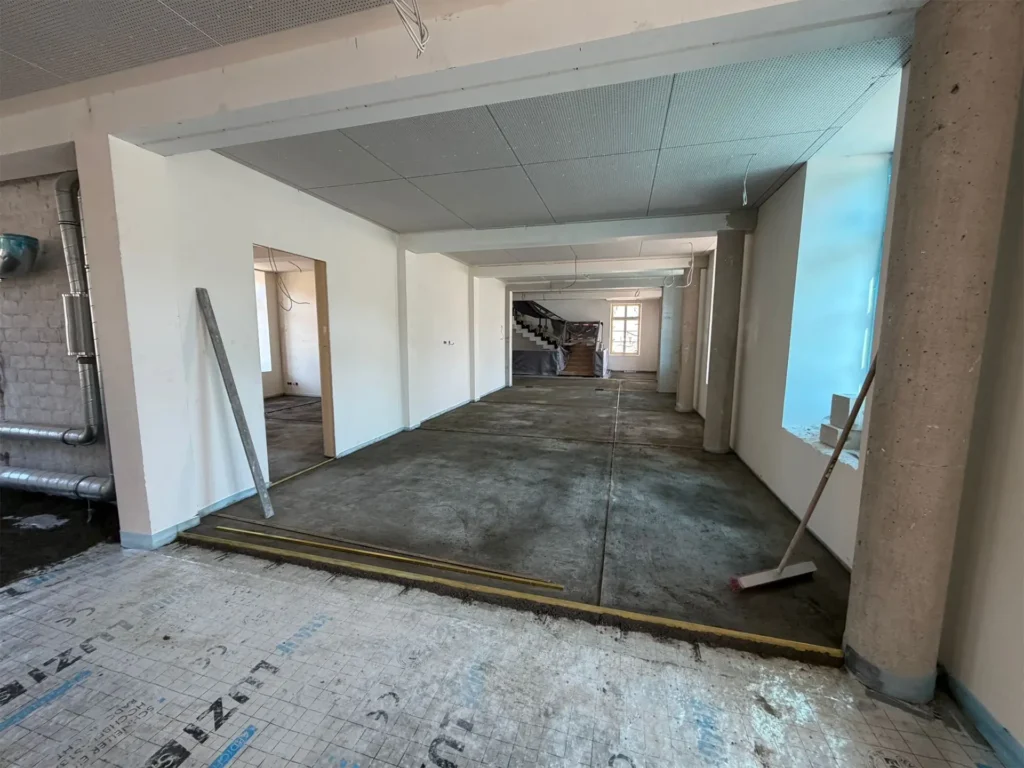 Heller, renovierter Innenraum im Umbau mit frisch verputzten weißen Wänden, rohem Betonboden und abgehängter Decke; im Hintergrund führt eine Treppe nach oben, rechts fällt Tageslicht durch ein Fenster, vereinzelt liegen Werkzeuge und Baumaterialien im Raum.