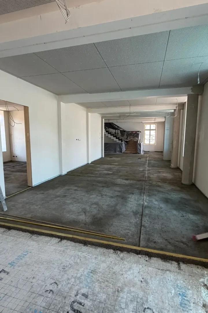 Heller, renovierter Innenraum im Umbau mit frisch verputzten weißen Wänden, rohem Betonboden und abgehängter Decke; im Hintergrund führt eine Treppe nach oben, rechts fällt Tageslicht durch ein Fenster, vereinzelt liegen Werkzeuge und Baumaterialien im Raum.