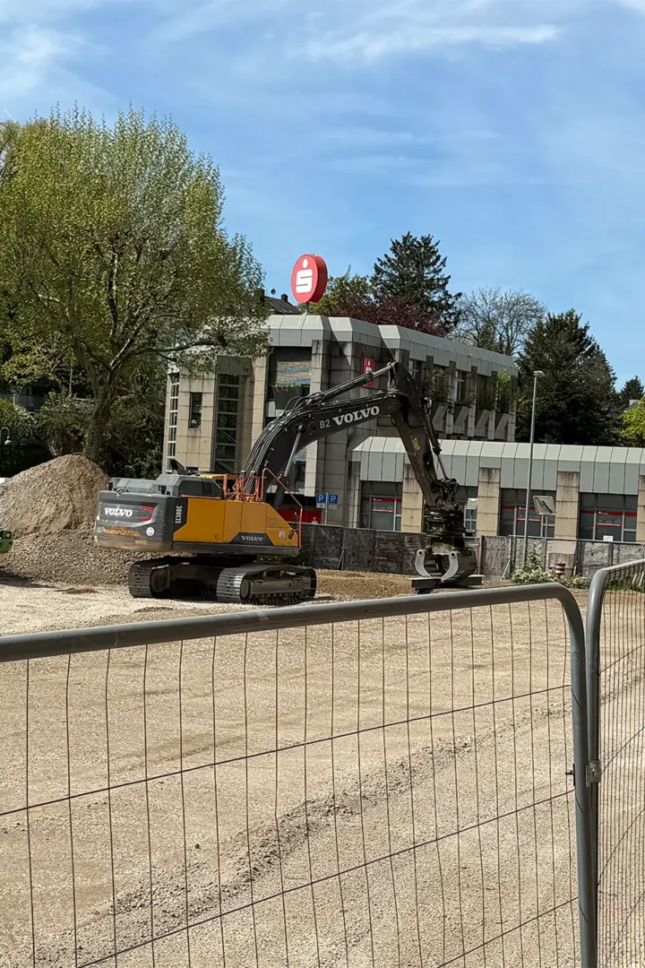 Baustelle vor einem Gebäude mit Sparkassen-Logo, auf der ein Bagger und ein Radlader Erdarbeiten durchführen, abgesperrt durch Bauzäune