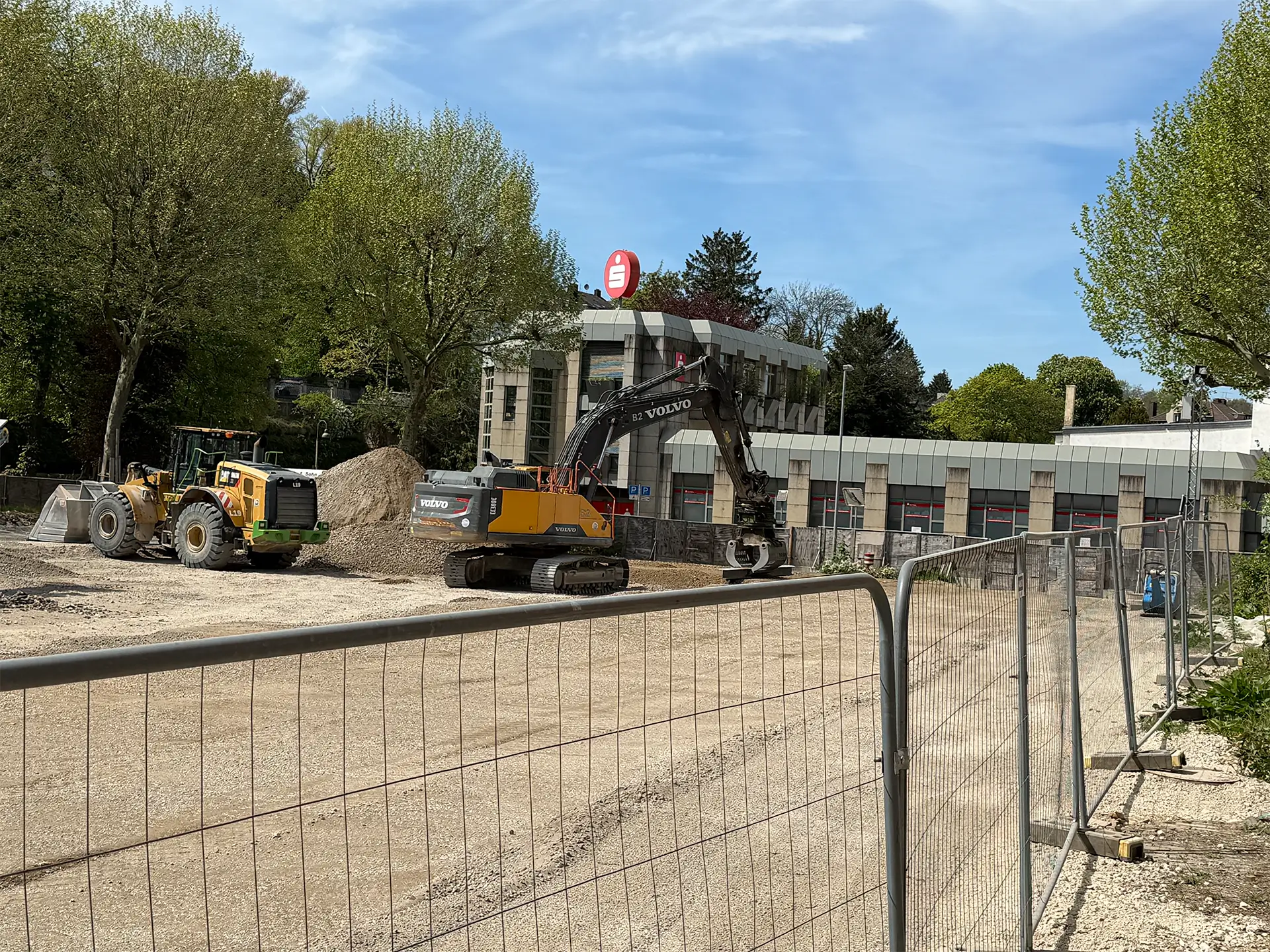 Baustelle vor einem Gebäude mit Sparkassen-Logo, auf der ein Bagger und ein Radlader Erdarbeiten durchführen, abgesperrt durch Bauzäune