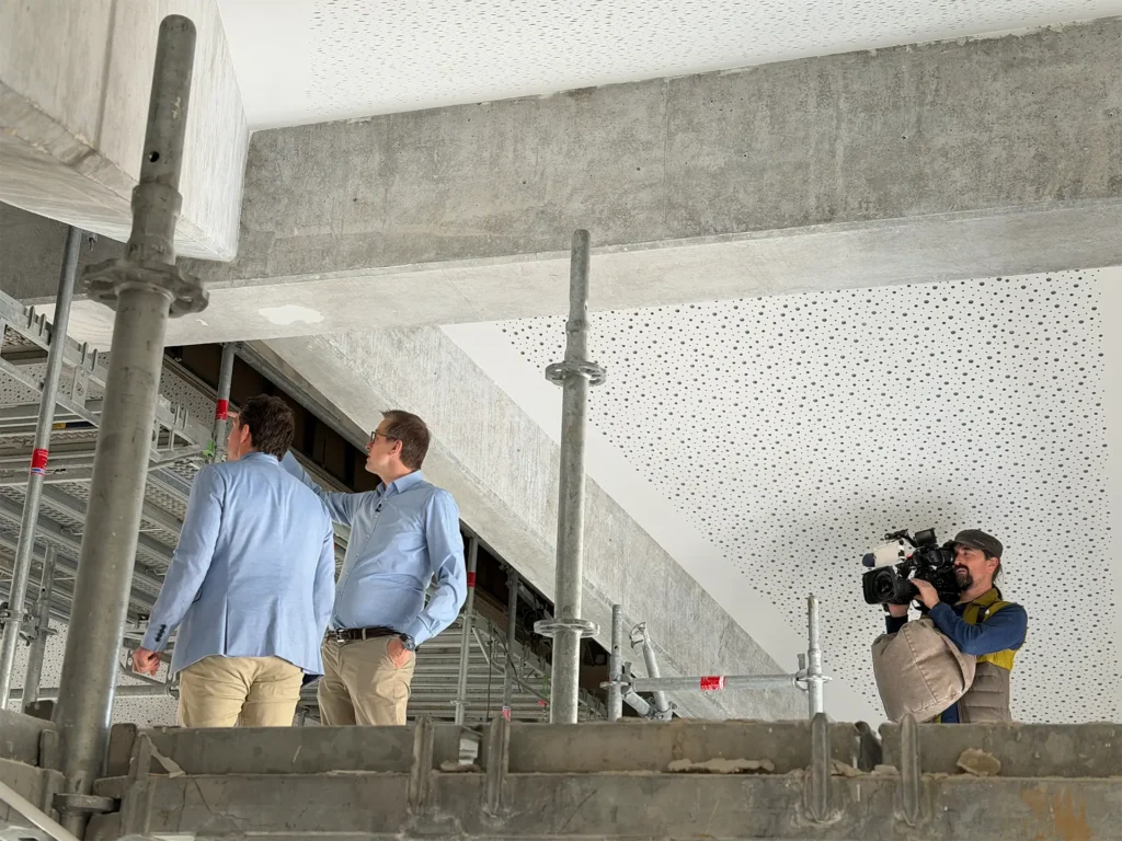 Zwei Männer besichtigen eine Baustelle im Innenbereich, während ein Kameramann die Szene filmt
