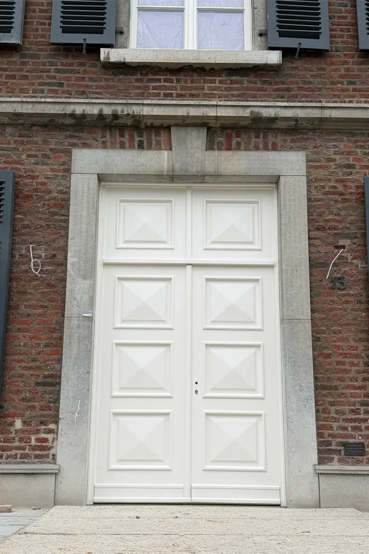 Historische Backsteinfassade eines sanierten Gebäudes mit großer weißer Eingangstür, hellen Natursteinrahmungen und dunkelgrauen Fensterläden.