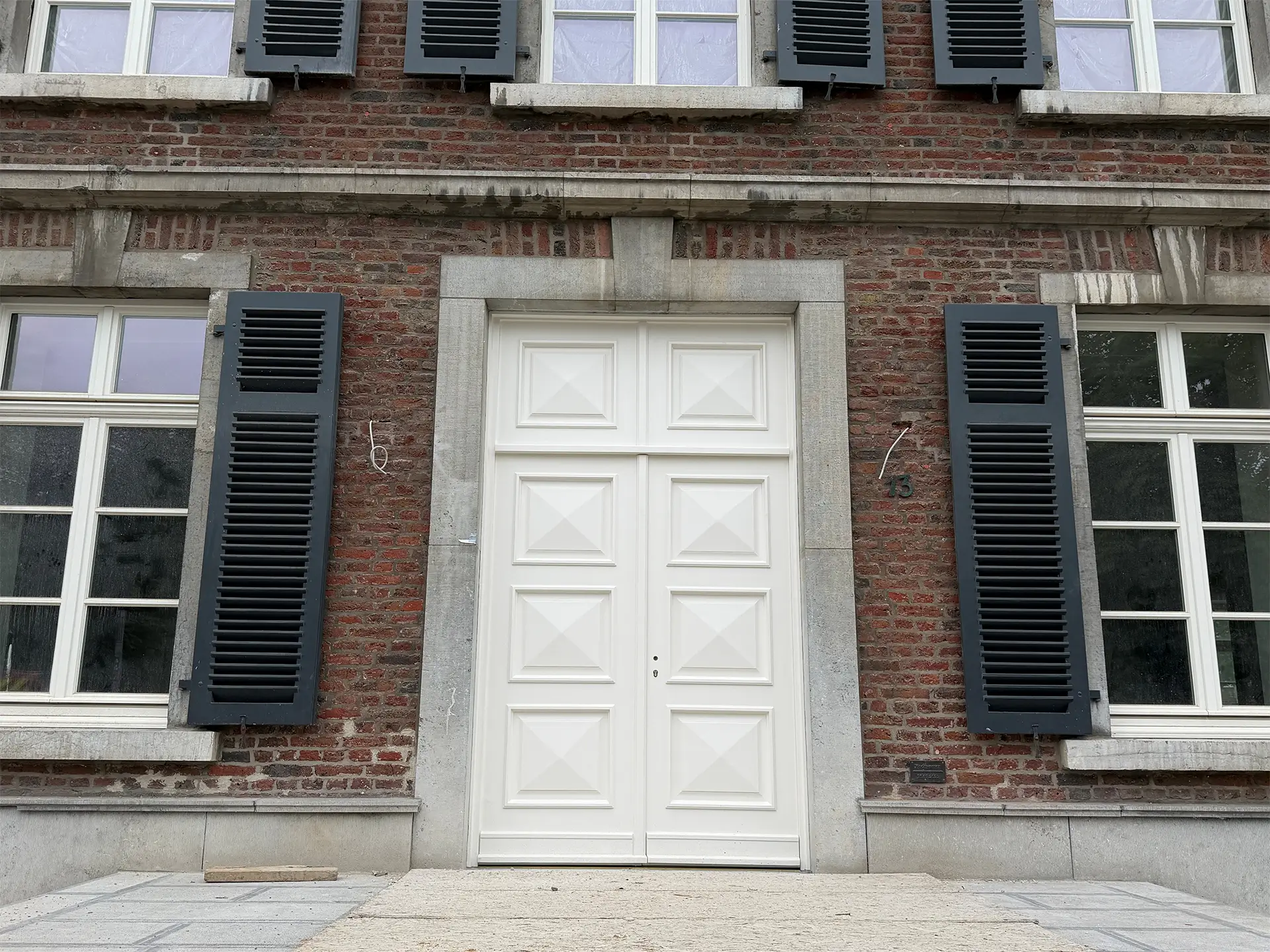 Historische Backsteinfassade eines sanierten Gebäudes mit großer weißer Eingangstür, hellen Natursteinrahmungen und dunkelgrauen Fensterläden.