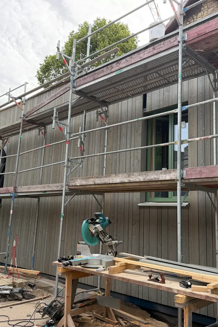 Gerüst und Kran vor einem Gebäude im Wiederaufbau. Die Holzfassade und grün gerahmte Fenster sind bereits montiert, während auf der Baustelle weitere Arbeiten an der Außenhülle erfolgen.