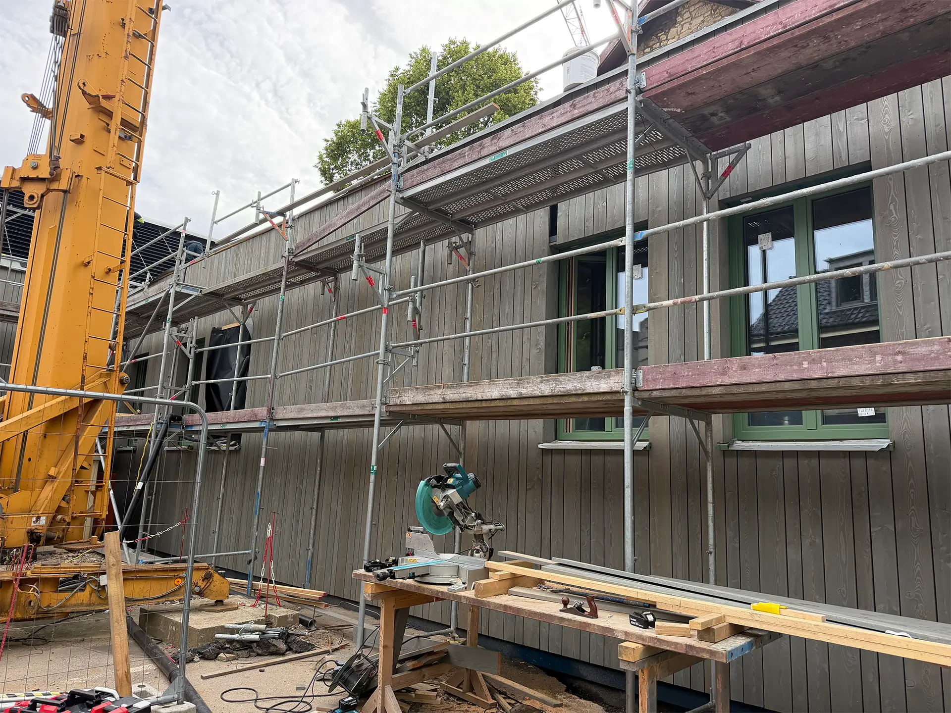 Gerüst und Kran vor einem Gebäude im Wiederaufbau. Die Holzfassade und grün gerahmte Fenster sind bereits montiert, während auf der Baustelle weitere Arbeiten an der Außenhülle erfolgen.