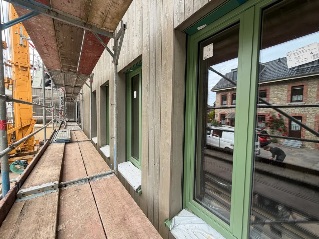 Blick entlang des Gerüsts auf die neu montierte Holzfassade mit grün gerahmten Fenstern. Die Fassadenarbeiten am Wiederaufbauprojekt in Stolberg schreiten sichtbar voran.