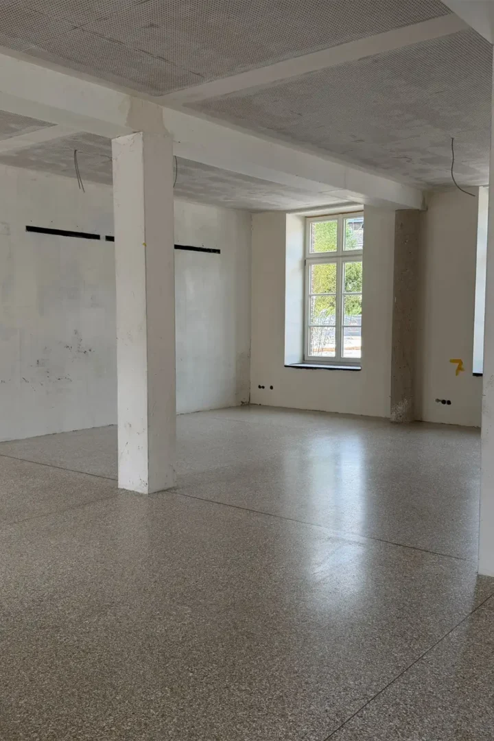 Heller, großzügiger Innenraum während der Sanierung mit frisch verlegtem Terrazzoboden, weißen Stützen und großen Fenstern mit Tageslichteinfall.