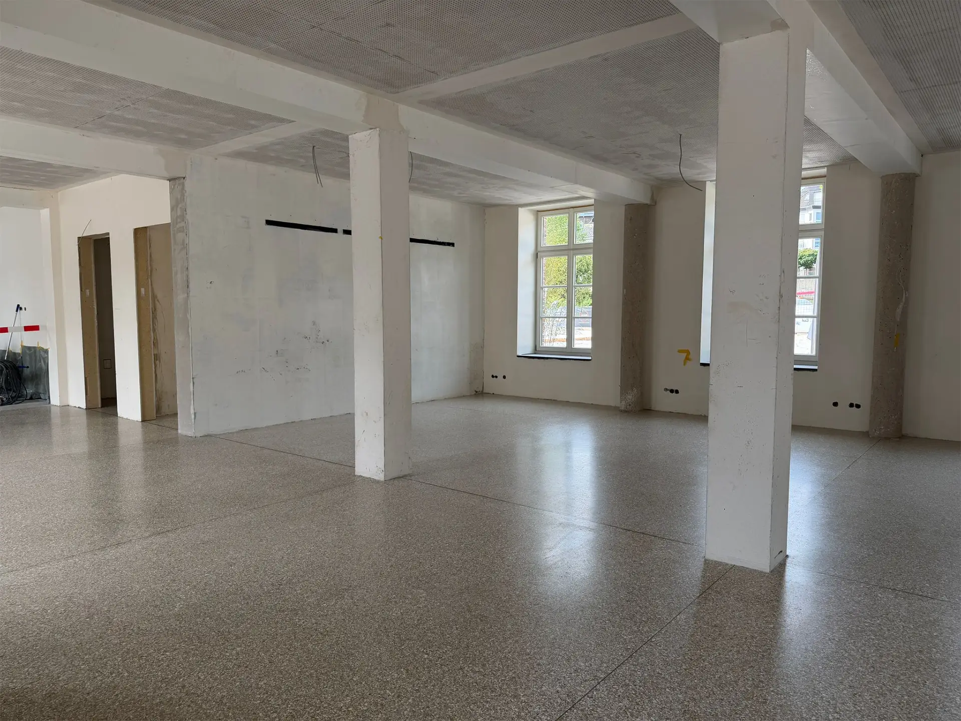 Heller, großzügiger Innenraum während der Sanierung mit frisch verlegtem Terrazzoboden, weißen Stützen und großen Fenstern mit Tageslichteinfall.