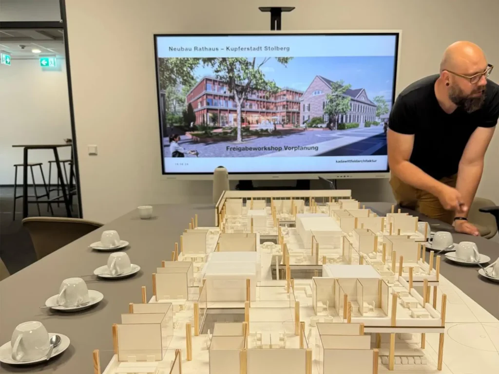 Architekt erläutert das Modell des geplanten Rathausneubaus Stolberg vor einer Visualisierung des Gebäudes während eines Planungsworkshops.