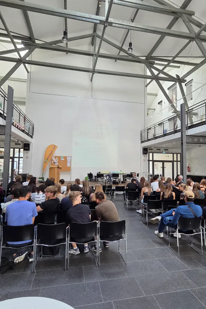 Blick in einen Veranstaltungsraum mit zahlreichen Teilnehmenden während einer Präsentation. Informationswände und Ausstellungselemente begleiten die Veranstaltung.