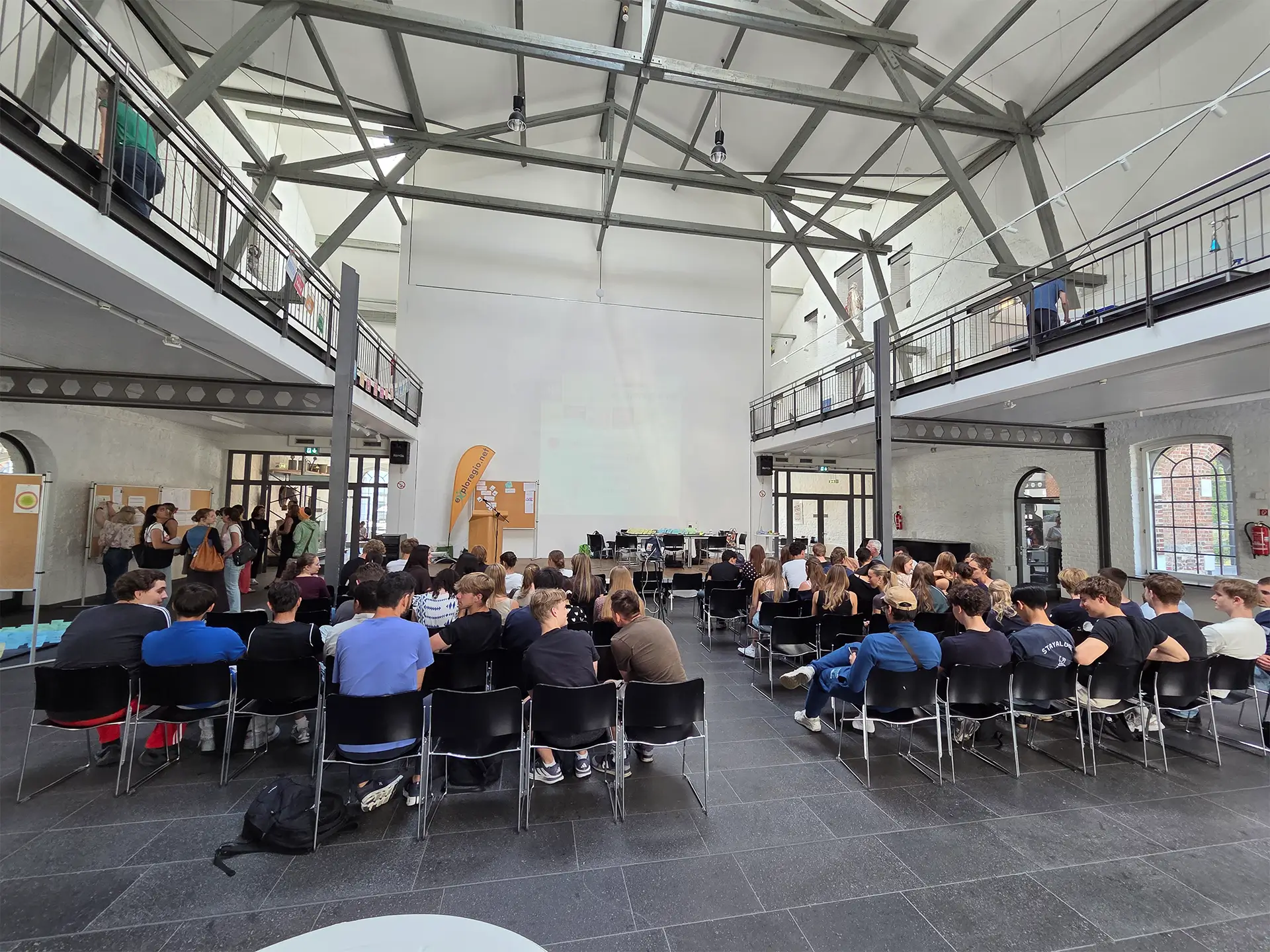 Blick in einen Veranstaltungsraum mit zahlreichen Teilnehmenden während einer Präsentation. Informationswände und Ausstellungselemente begleiten die Veranstaltung.