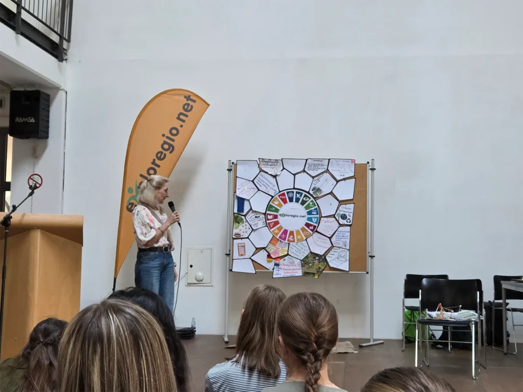 Eine Referentin präsentiert die Ergebnisse eines Workshops vor einer Pinnwand mit Ideen und Notizen. Im Vordergrund verfolgen die Teilnehmenden den Vortrag.