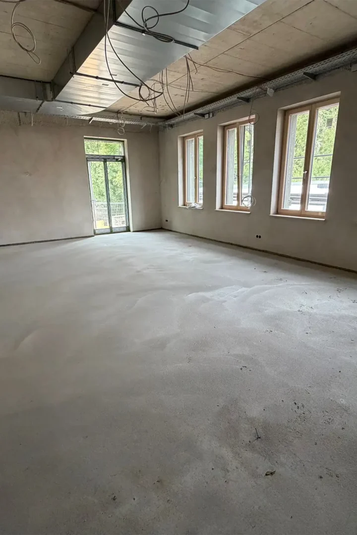Großer, heller Innenraum im Rohbau des neuen Rathauses in Stolberg mit frisch verputzten Wänden, Estrichboden, sichtbaren Lüftungskanälen und Elektroinstallationen an der Decke sowie einer Fensterfront mit Blick nach draußen.