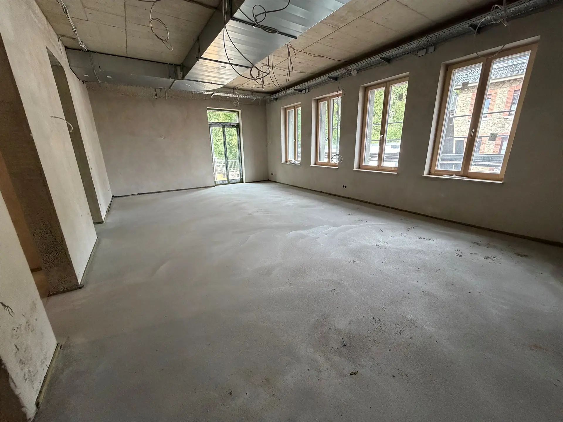 Großer, heller Innenraum im Rohbau des neuen Rathauses in Stolberg mit frisch verputzten Wänden, Estrichboden, sichtbaren Lüftungskanälen und Elektroinstallationen an der Decke sowie einer Fensterfront mit Blick nach draußen.