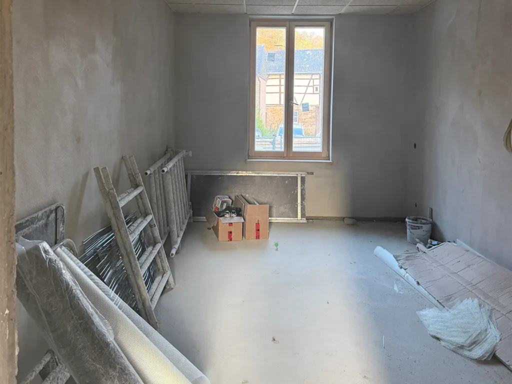Sanierungsarbeiten in einem Raum der Kita Vicht mit großen Fenstern, verputzten Wänden und Baumaterialien.